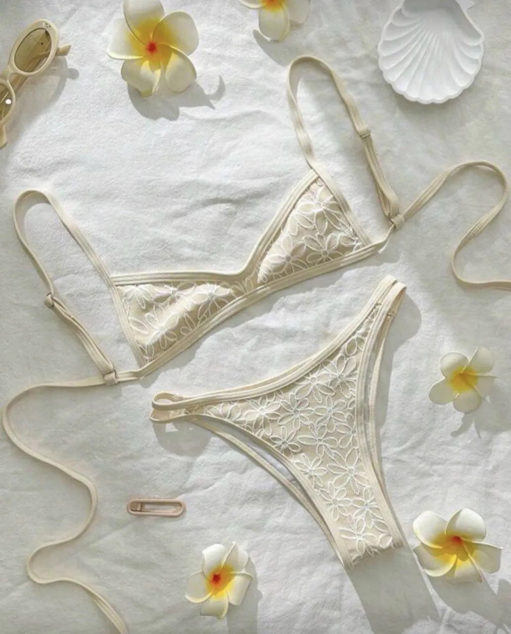 Bikini de dos piezas color crudo con detalles de flores blancas bordadas. La parte superior es un corpiño triangular con tiras finas regulables. La parte inferior es un culotte con tiras finas a los costados.