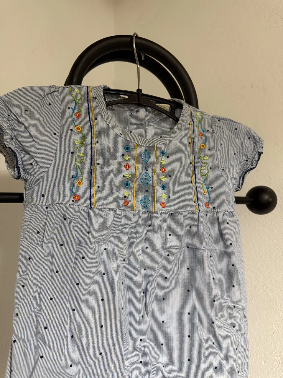 Remera a rayas celestes con lunares negros y bordado floral en el frente. Tiene cuello redondo y mangas cortas con terminación fruncida.