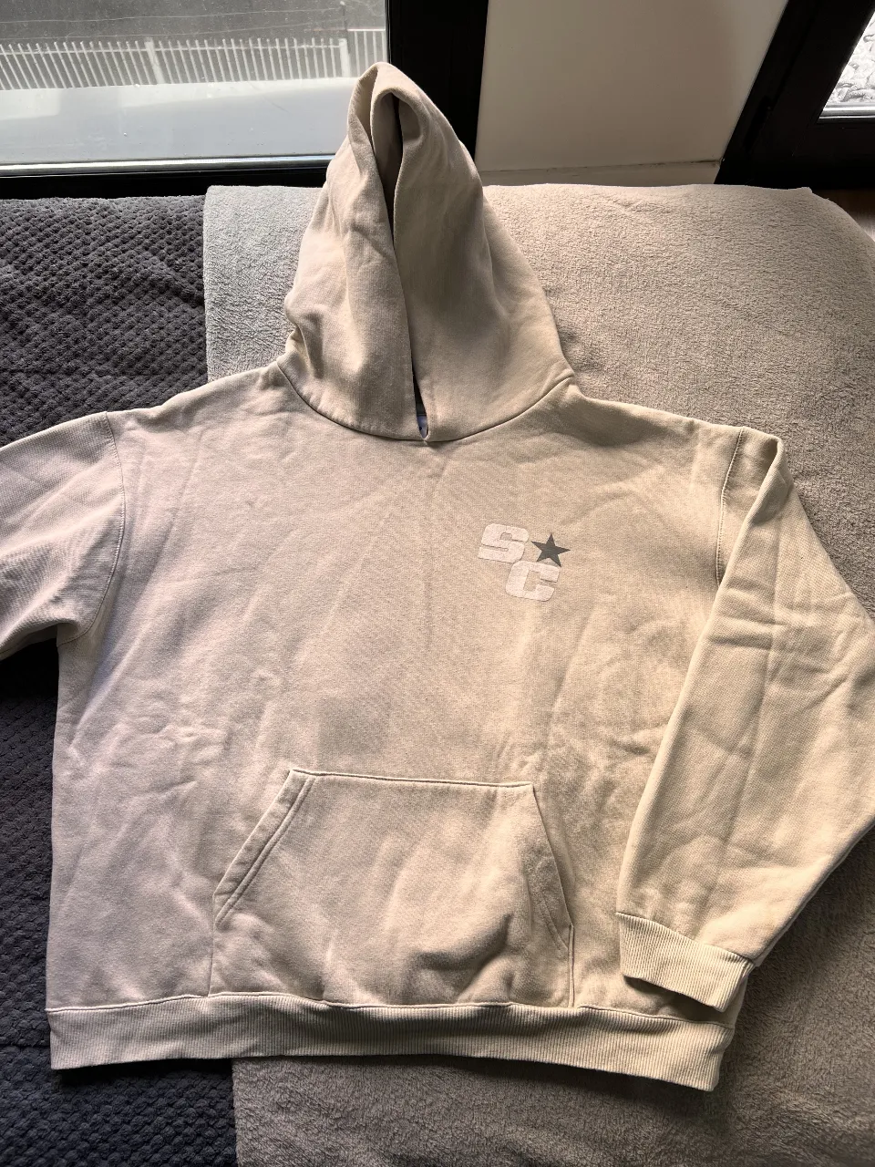 Buzo canguro de color crudo con capucha. En el frente, un logo discreto y en la espalda, un diseño con texto y estrellas. Ideal para un look casual y cómodo.