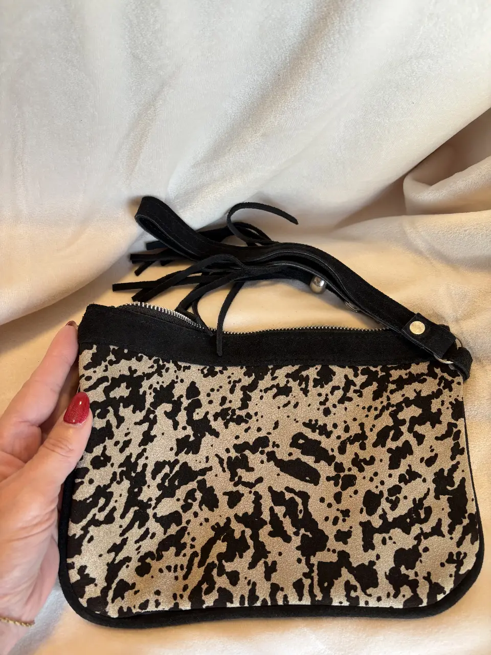 Cartera de mano con estampado animal print en tonos crudo y negro. Posee un detalle de flecos y una correa para llevar en la muñeca. Ideal para una salida.