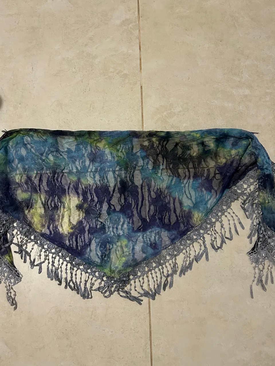 Pañuelo triangular con un estampado abstracto en tonos azules, verdes y amarillos. Posee un delicado borde de flecos plateados que le dan un toque bohemio y chic. Ideal para complementar outfits de primavera o verano.