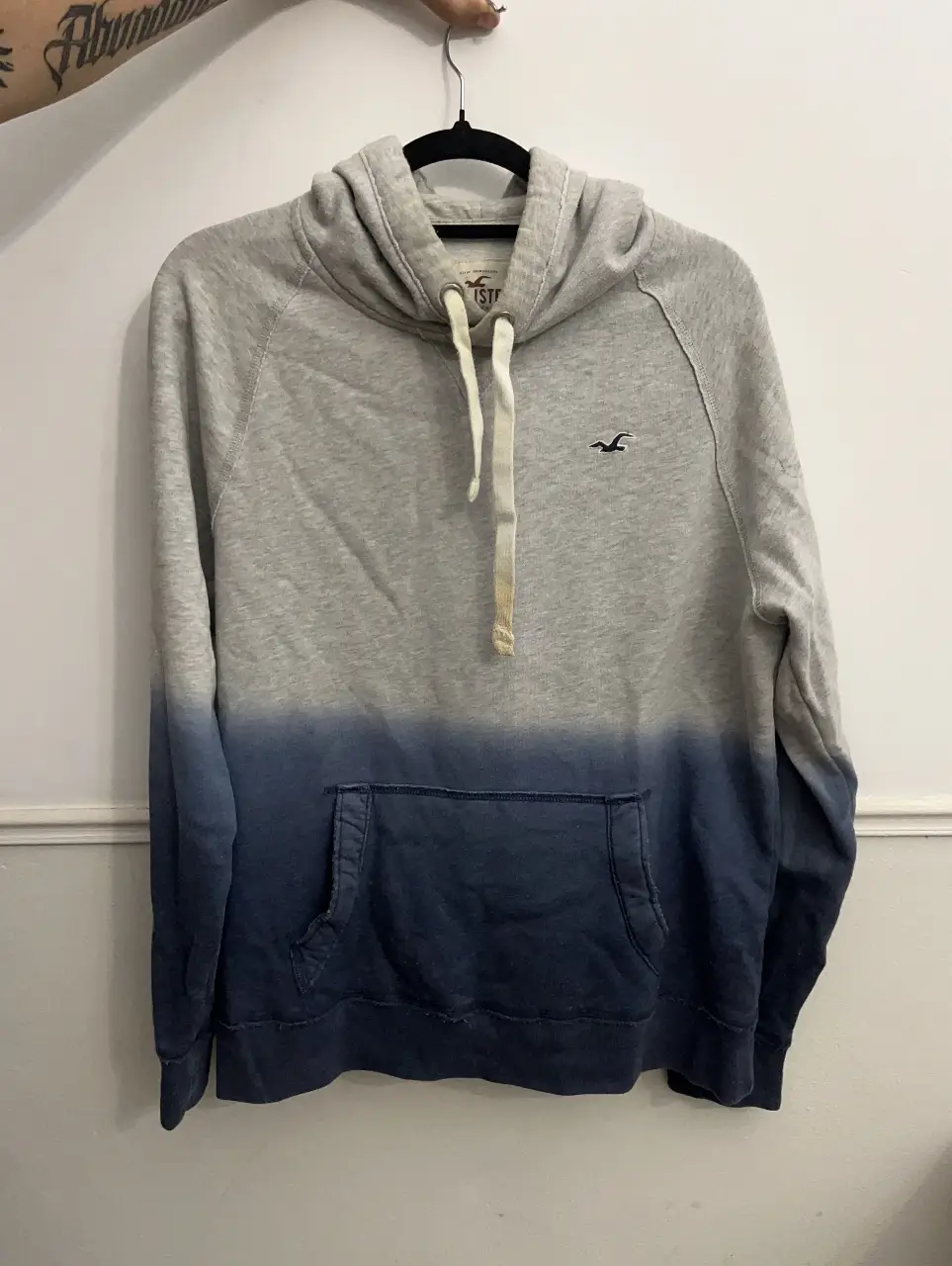 Buzo canguro Hollister con capucha, de color gris con degradado a azul en la parte inferior. Ideal para un look casual y cómodo.