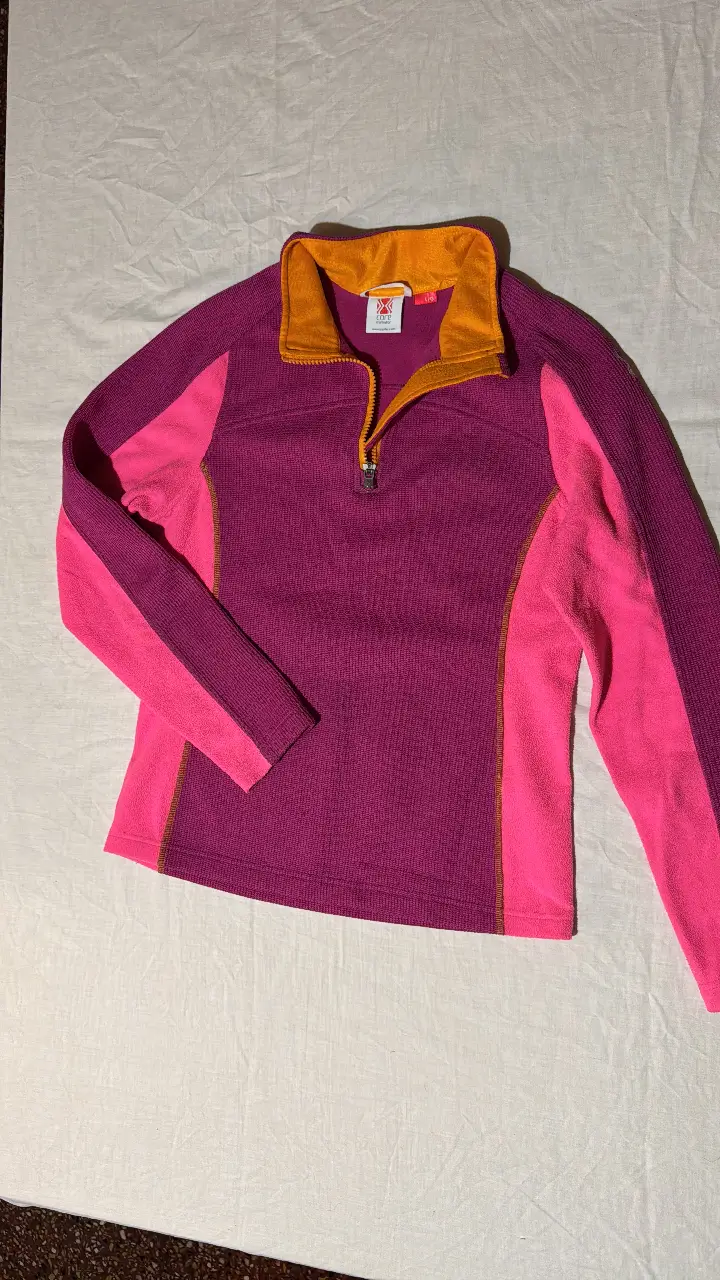 Buzo con cierre y cuello alto en tonos rosa y violeta. Detalles en naranja y cierre metálico. Ideal para actividades al aire libre.