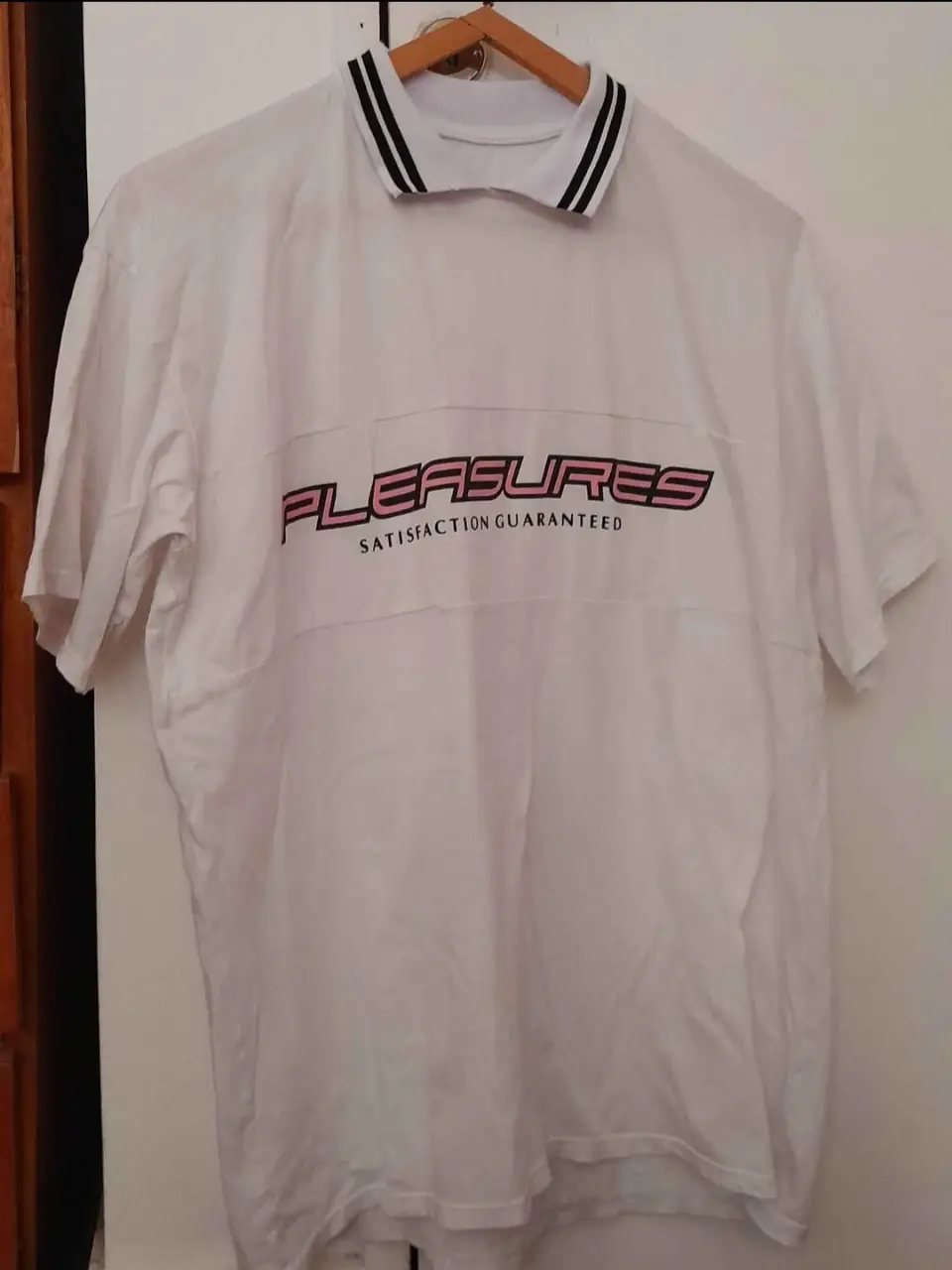 Remera blanca con cuello polo y detalles en rosa. Estampado "PLEASURES SATISFACTION GUARANTEED". Ideal para un look casual y moderno.