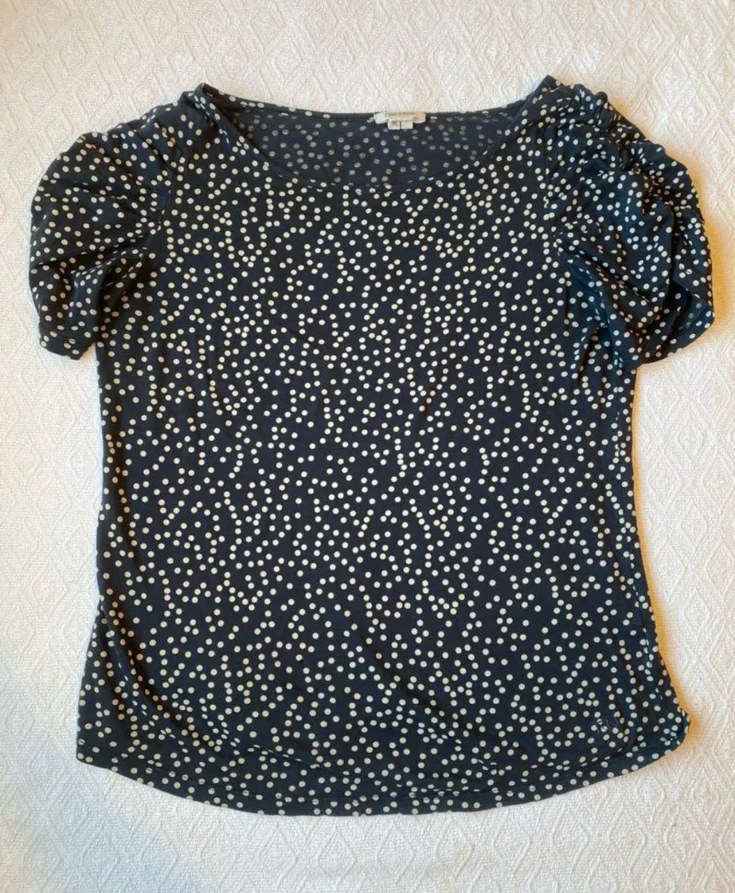 Blusa de mujer con estampado de lunares blancos sobre fondo azul marino. Confeccionada en 100% algodón Pima, manga corta con detalle fruncido en los hombros. Ideal para un look casual y cómodo.