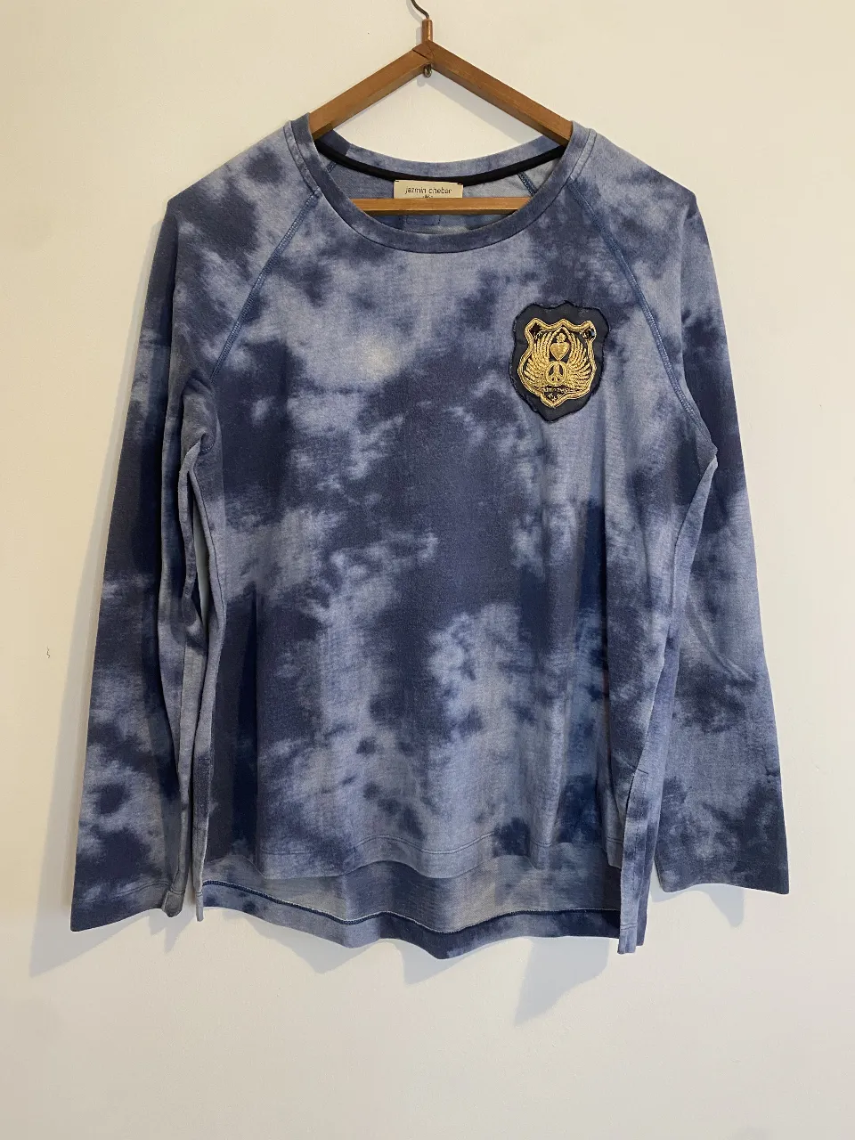 Buzo de Jazmín con diseño tie-dye en tonos azules y detalle de parche dorado en el pecho. Ideal para un look casual y moderno. Sólo tiene el detalle de un poco raspado en el pecho 