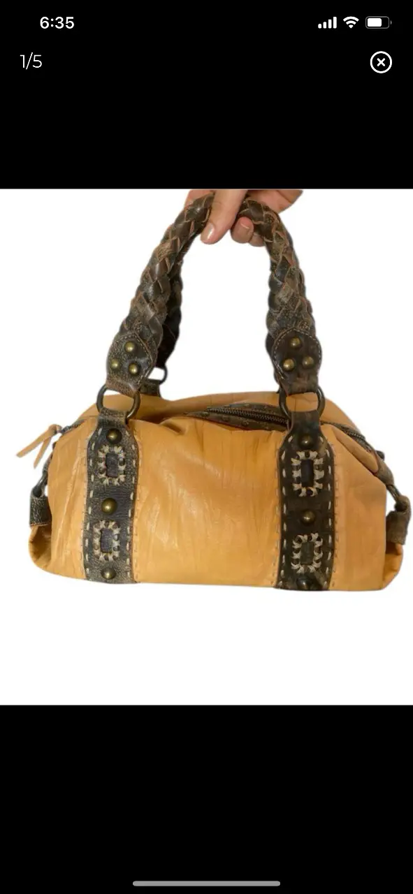 Cartera de mano con detalles artesanales. Diseño en cuero color mostaza con detalles en marrón y apliques metálicos. Manijas trenzadas y cierre superior. Ideal para un look bohemio y con estilo. 33x20cm