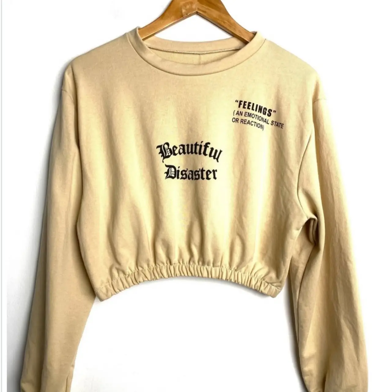 Buzo crop de color crudo con estampado "Beautiful Disaster" en la parte delantera. Tiene mangas largas y un ajuste elástico en la cintura. En la parte superior derecha, tiene la frase "FEELINGS" (AN EMOTIONAL STATE OR REACTION). Ideal para un look casual y moderno. hombros 49 mangas 56 largo 40