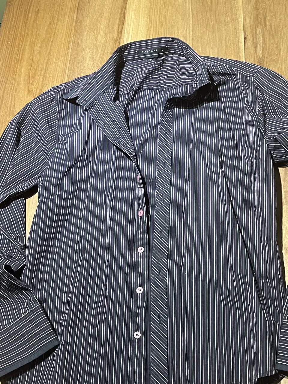 Camisa de hombre a rayas azul y blanca, manga larga, con botones rosas y etiqueta de la marca Tascani. Ideal para un look casual o de trabajo.