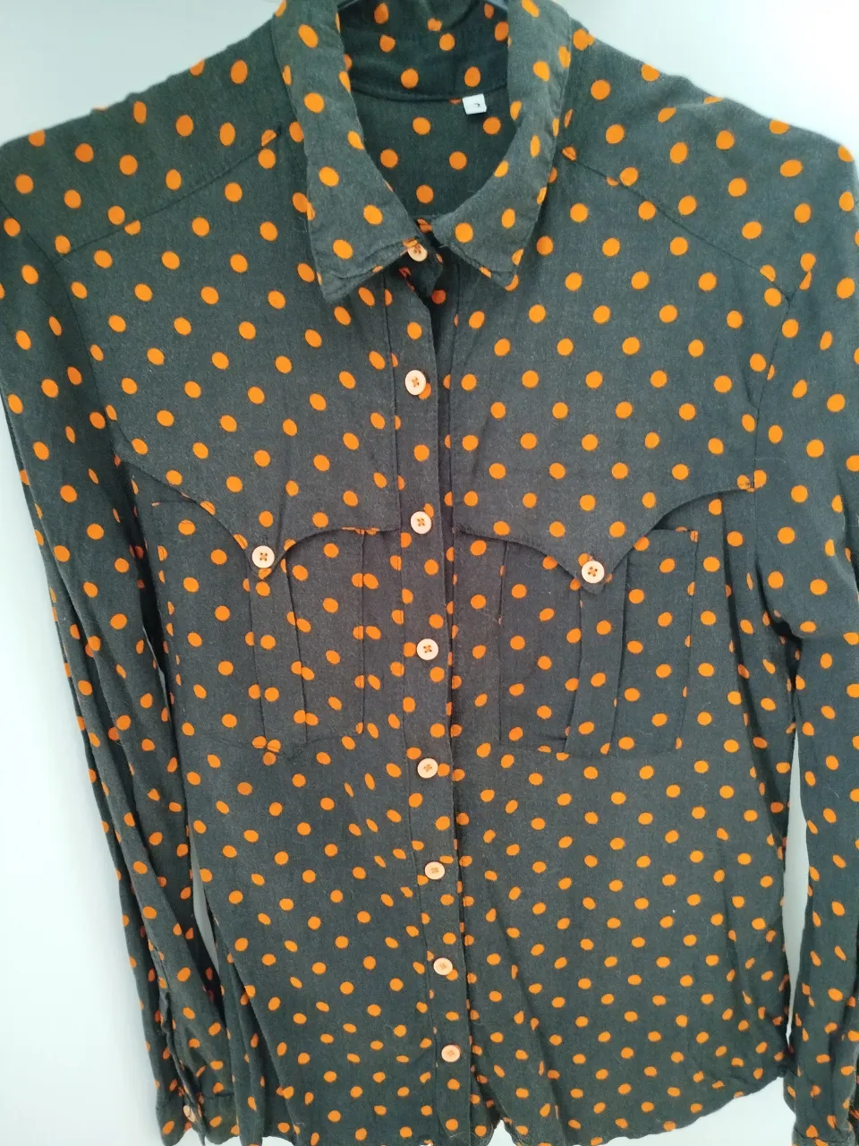Camisa de mujer con estampado de lunares naranjas sobre fondo negro. Diseño clásico con botones y bolsillos en el pecho. Ideal para un look casual con un toque retro.