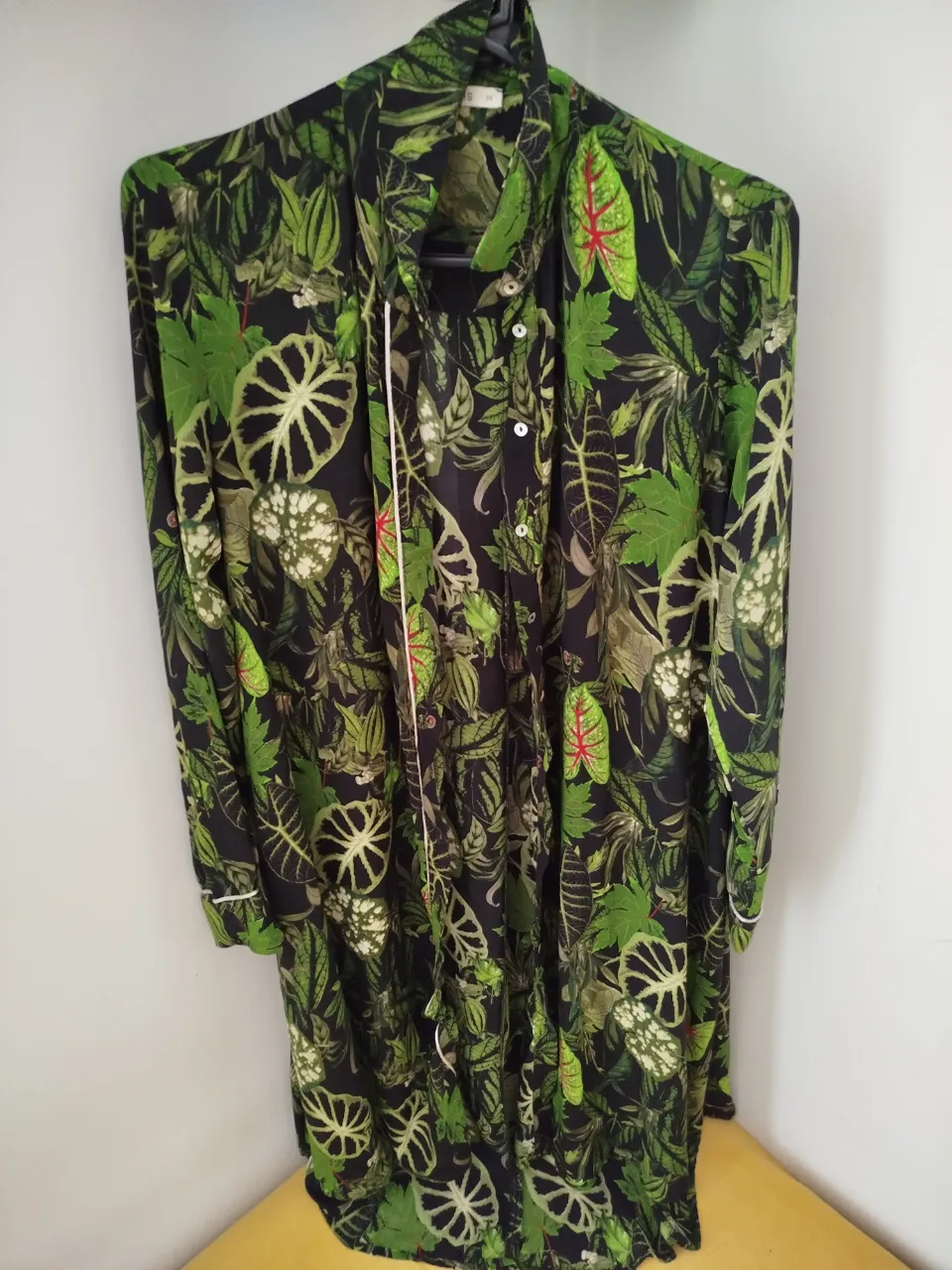 Camisa estampada con hojas tropicales en tonos verdes y negros. Diseño con botones y detalle de cinta blanca. Ideal para un look fresco y moderno.