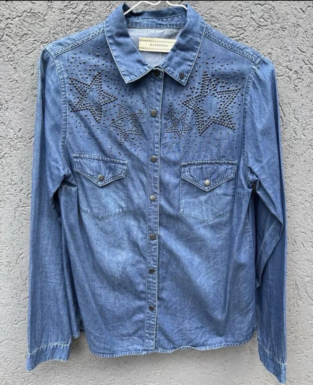 Camisa de jean azul con tachas y estrellas bordadas en el frente. Tiene bolsillos con solapa y botones metálicos. Un diseño único y canchero, ideal para darle un toque rockero a tu look.