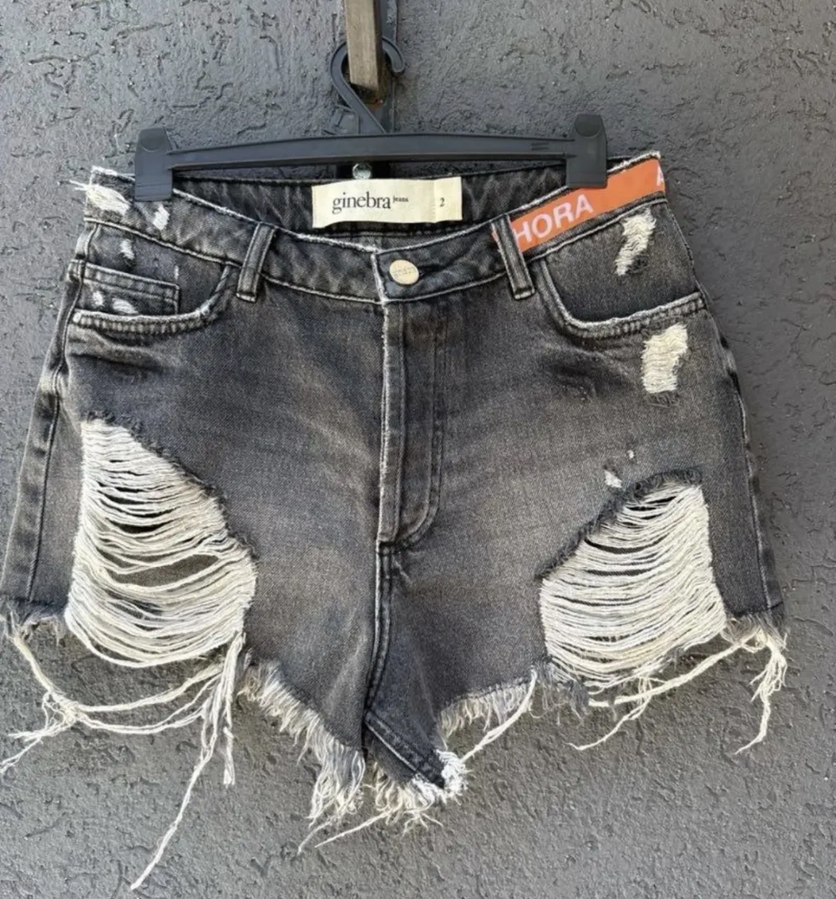 Shorts de jean grises  roturas y detalles deshilachados. Tiro alto con cinta naranja con la palabra "AHORA" estampada. Perfectos para un look canchero y moderno.