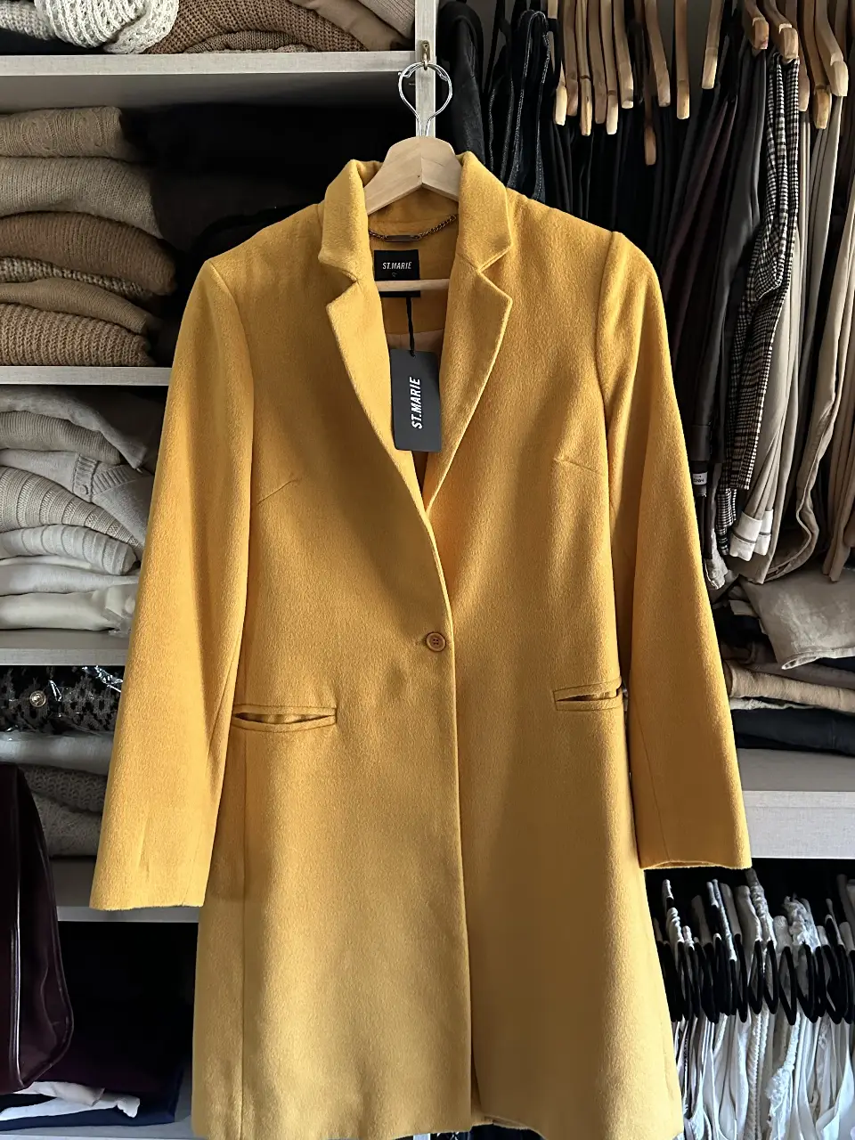 Elegante tapado largo de color amarillo mostaza, ideal para un look formal o de trabajo. Confeccionado en paño, este blazer de la marca St. Marie presenta un diseño clásico con solapas y un solo botón.