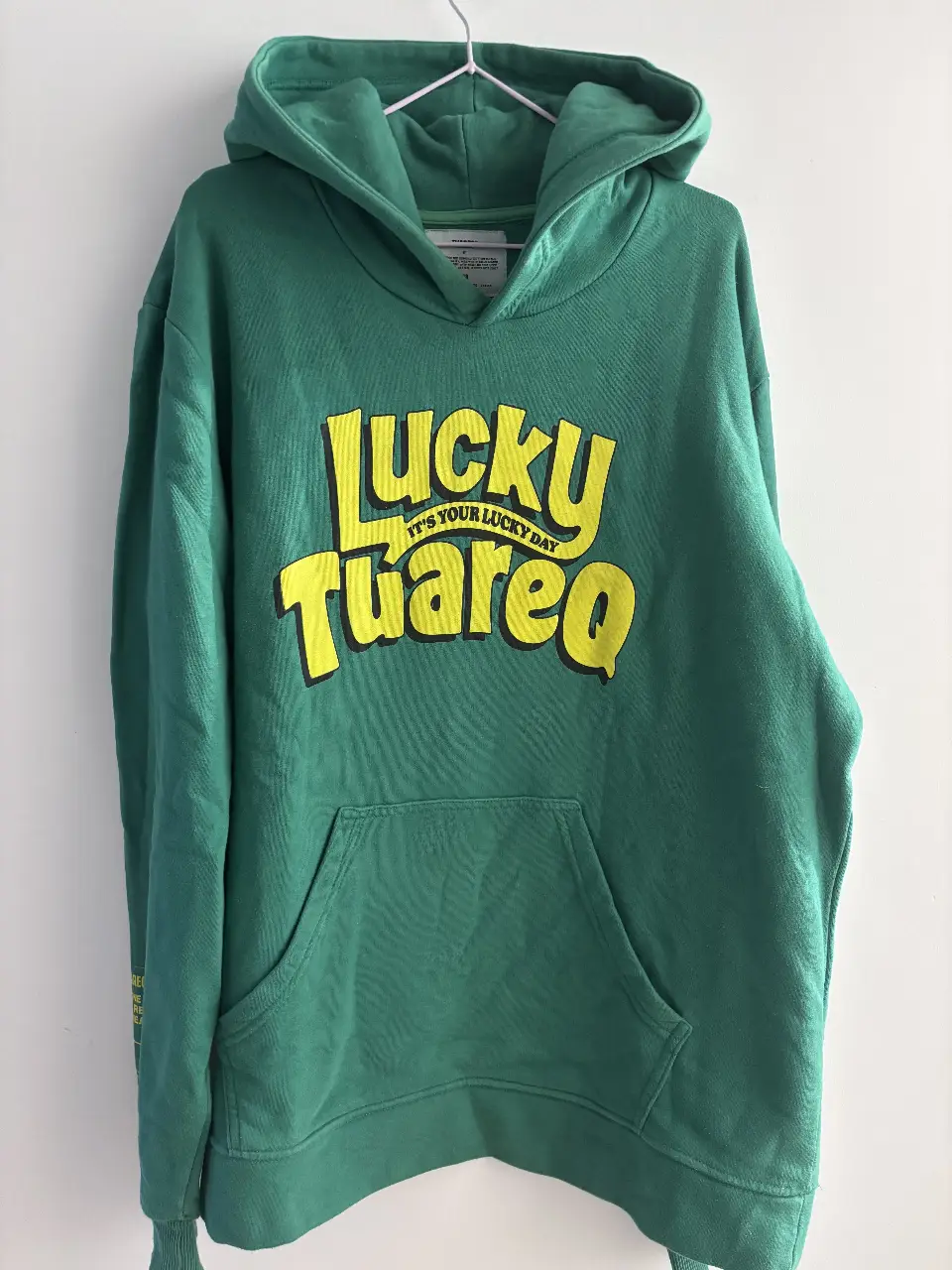 Buzo canguro verde con estampado "LUCKY TUAREQ" en amarillo. En la espalda, frase "Look For Something Positive In Each Day". Detalles de la marca en la manga y etiqueta en el interior. Ideal para un look casual y cómodo.