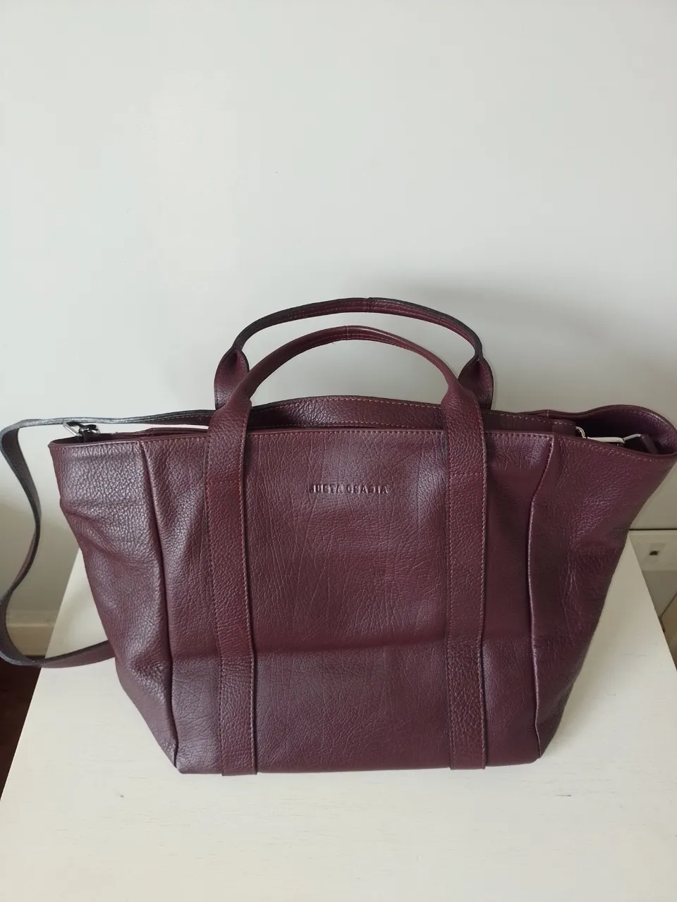 Bolso de cuero color bordó, súper práctico y canchero. Tiene dos asas cortas y una correa larga desmontable para llevar cruzado o al hombro. El interior es amplio y tiene bolsillos para organizar todo. ¡Un básico que no te puede faltar!