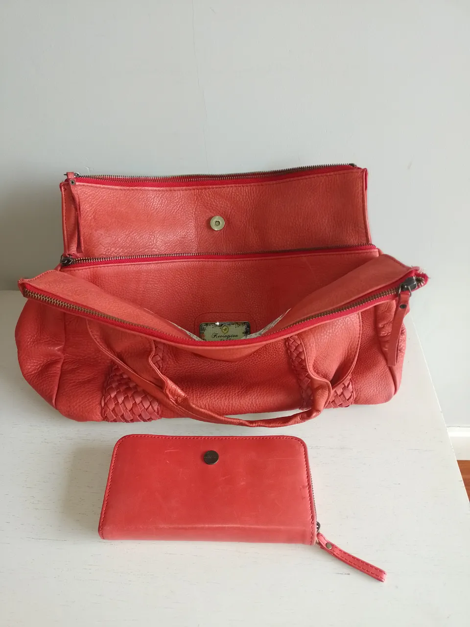 Set de cartera y billetera de cuero color coral. La cartera tiene manijas cortas y un diseño tejido en los laterales. La billetera es rectangular y se cierra con un broche.