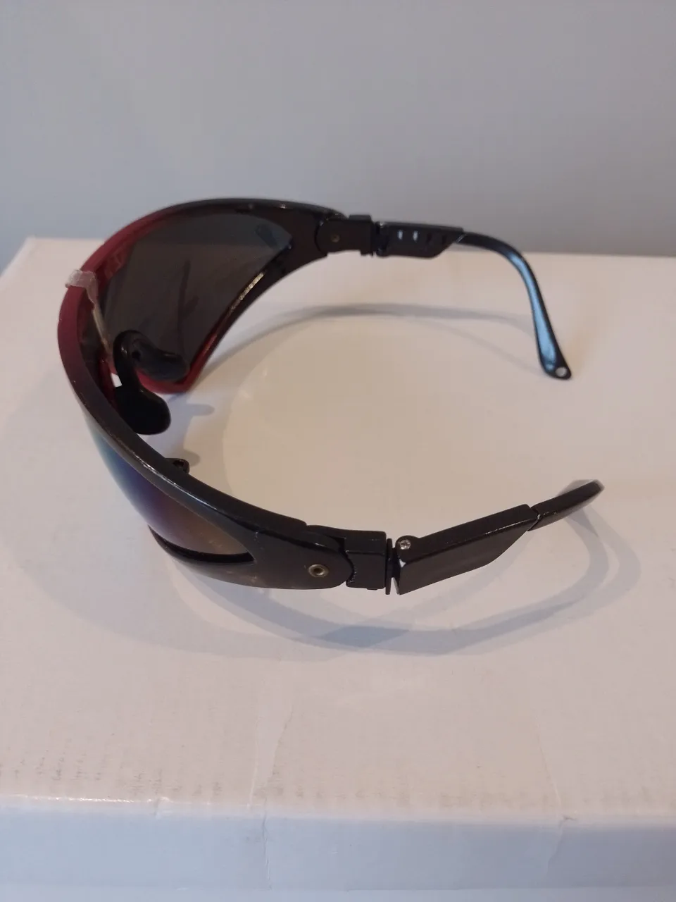 Anteojos deportivos con marco negro y detalles en rojo. Lentes espejados con reflejos azules y amarillos. Marca "THE GAP" visible en el frente.