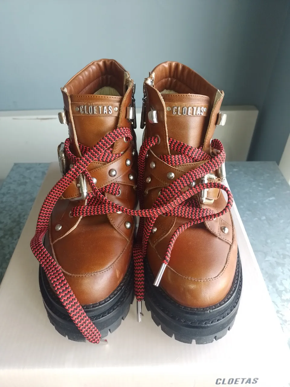 Botitas de cuero marrón con cordones rojos y negros, detalles metálicos y cierre lateral. Suela gruesa y robusta, ideales para un look urbano y canchero.