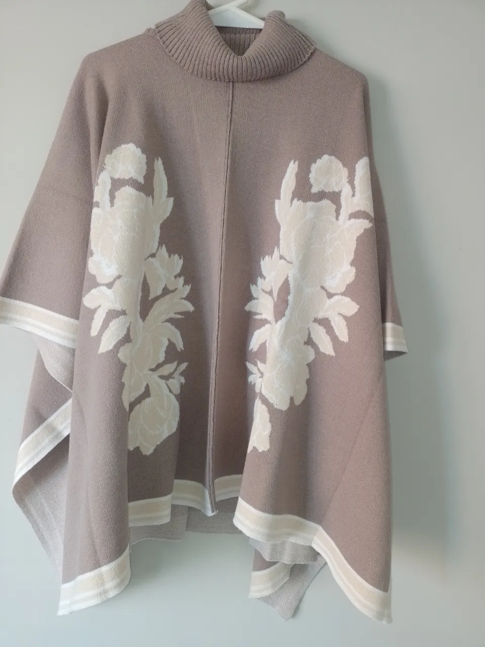 Poncho de tejido color tiza con cuello alto y estampa floral en blanco y crudo. Posee detalles de tiras a rayas en los bordes inferiores y en las mangas.