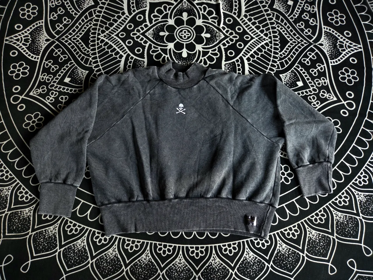 Buzi gris oscuro estilo gastaso, con cuello alto y bordado de calavera con tibias cruzadas en el frente. Posee puños y cintura elastizados. Es corto, mangas murcielago, oversize