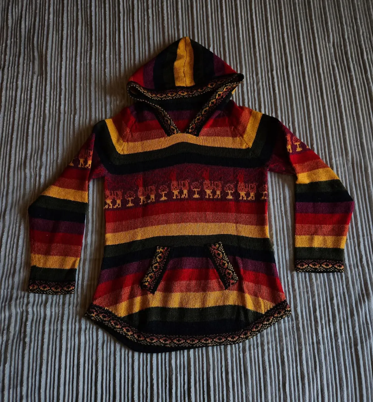 Buzo tejido de alpaca con capucha y bolsillos frontales. Presenta un diseño de rayas horizontales multicolor y un estampado de figuras de animales en la parte superior del torso. Los puños y el borde inferior tienen un tejido decorativo.