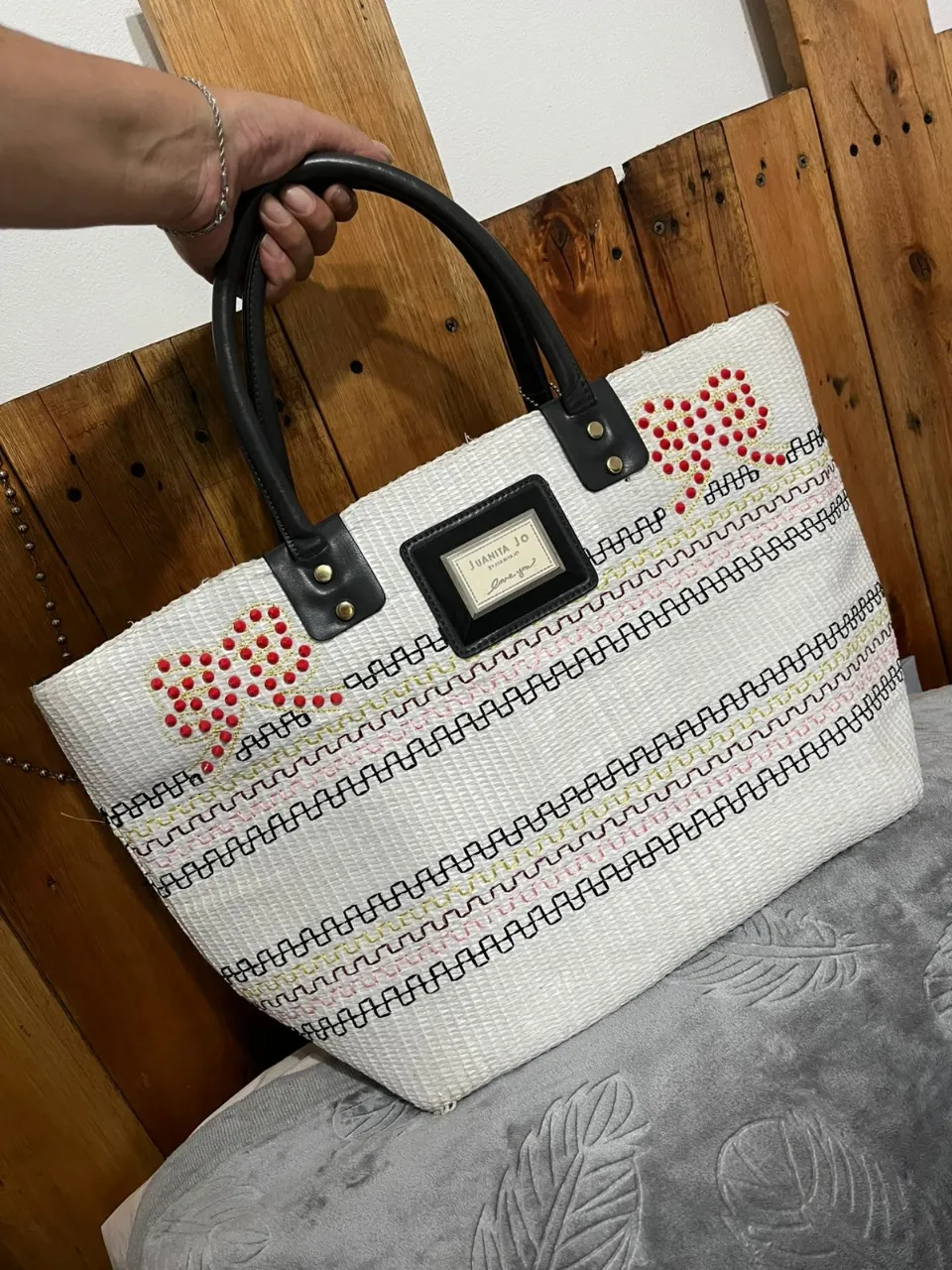 Bolso blanco tejido con manijas negras. Posee detalles bordados en forma de moños y líneas horizontales de colores rosa, verde y negro. Tiene una etiqueta negra con el logo de la marca.