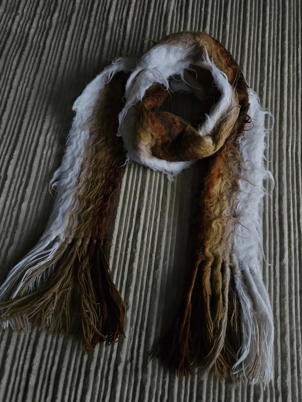 Bufanda tejida con flecos largos en los extremos. Presenta un diseño de franjas verticales en tonos marrones, blancos y ocres.