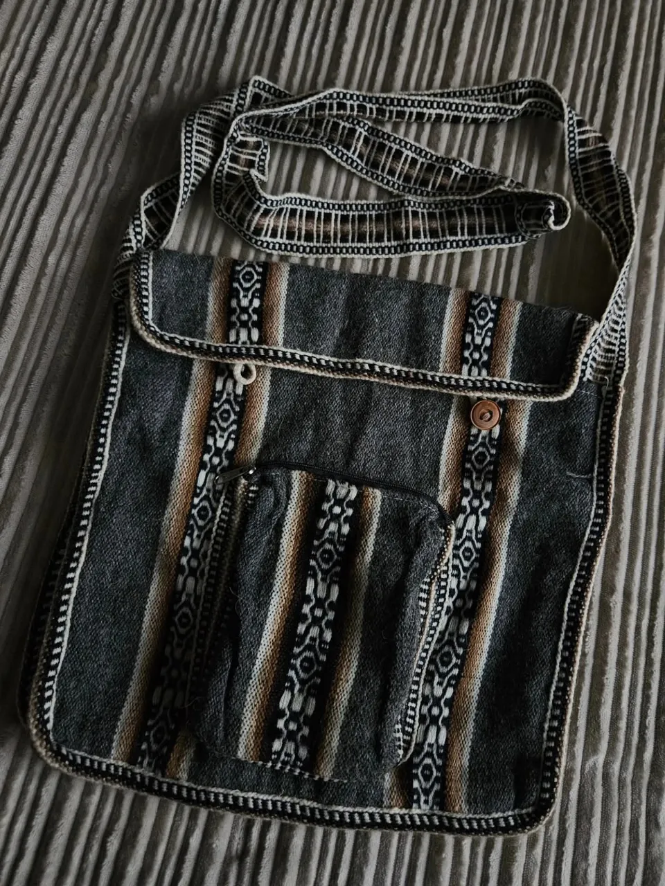 Bolso de tela con correa regulable. Posee un bolsillo frontal con cierre y otro bolsillo con botón a presión. Diseño de rayas verticales en tonos grises, marrones y blancos con detalles de guardas tejidas.