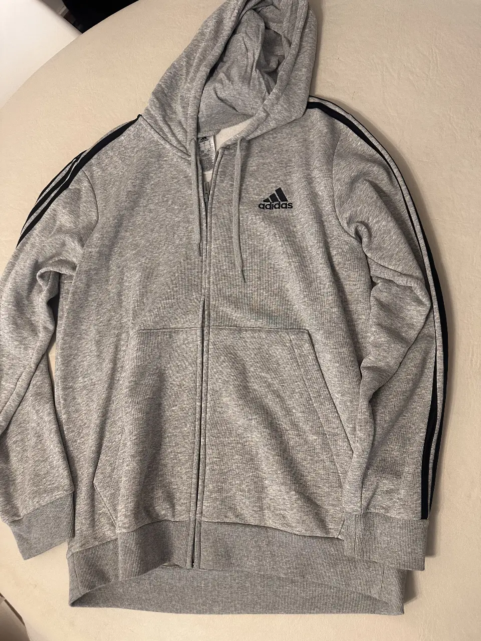 Buzo Adidas con cierre, color gris con detalles en negro. Ideal para un look deportivo y casual. Cuenta con el logo de la marca y las clásicas tres tiras en las mangas.