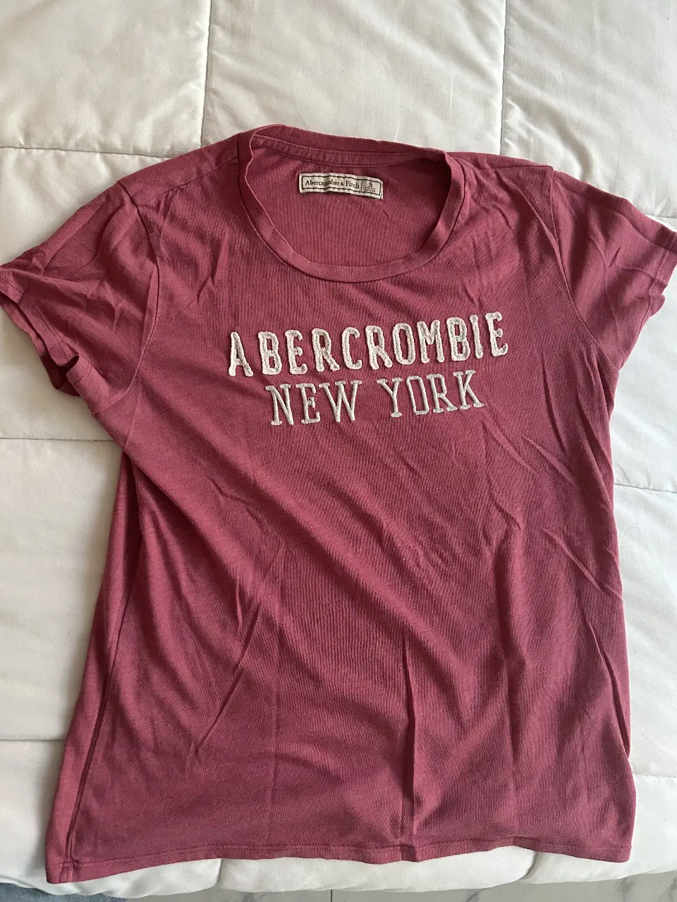Remera de manga corta Abercrombie & Fitch, color rosa viejo con estampado "Abercrombie New York" en blanco. Ideal para un look casual y cómodo.