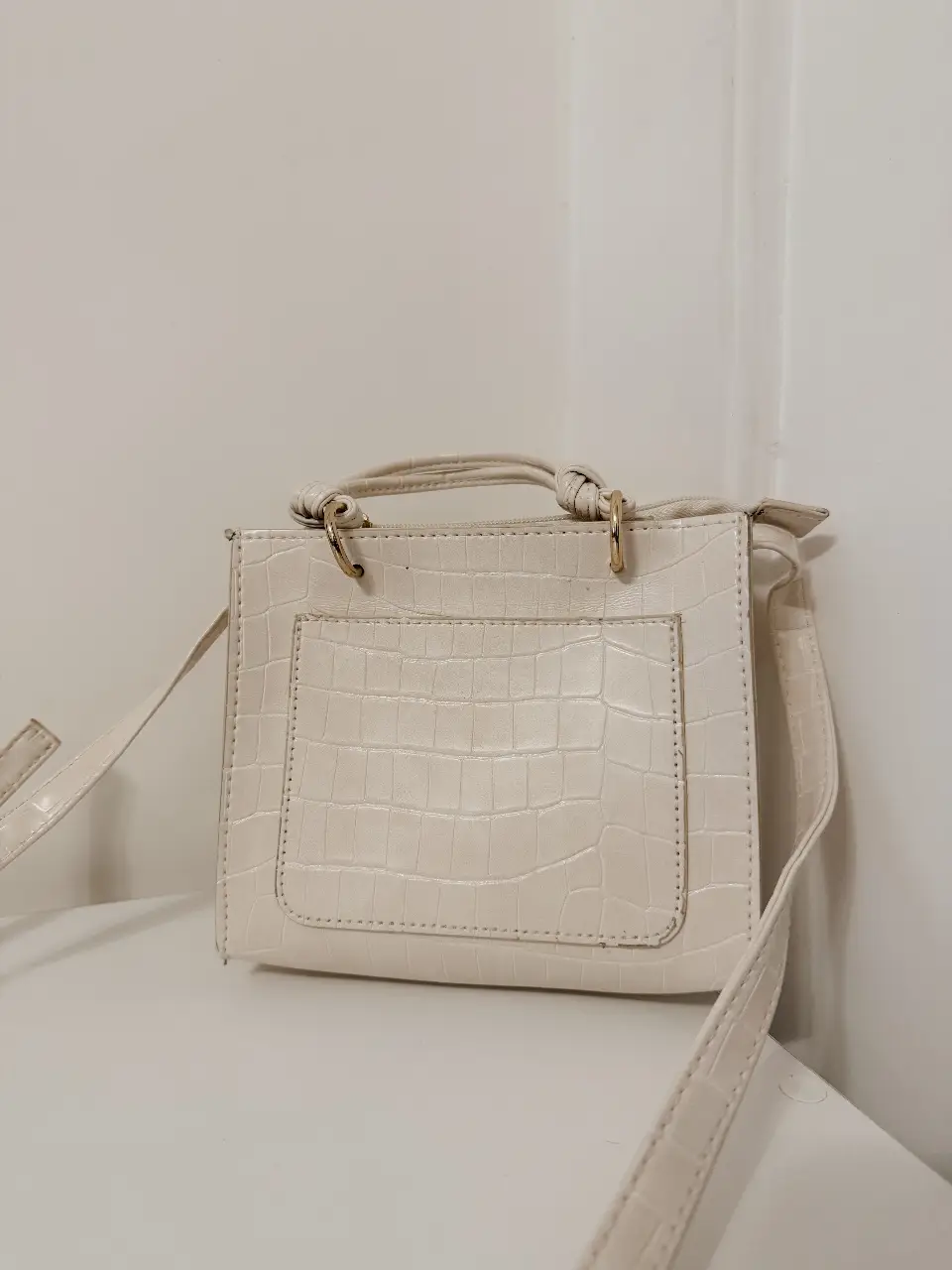 Cartera bandolera de color blanco, con textura símil cuero de cocodrilo. Cuenta con manijas cortas y correa larga ajustable. En su interior, se encuentra una etiqueta con la marca Primark. Ideal para un look casual y moderno.