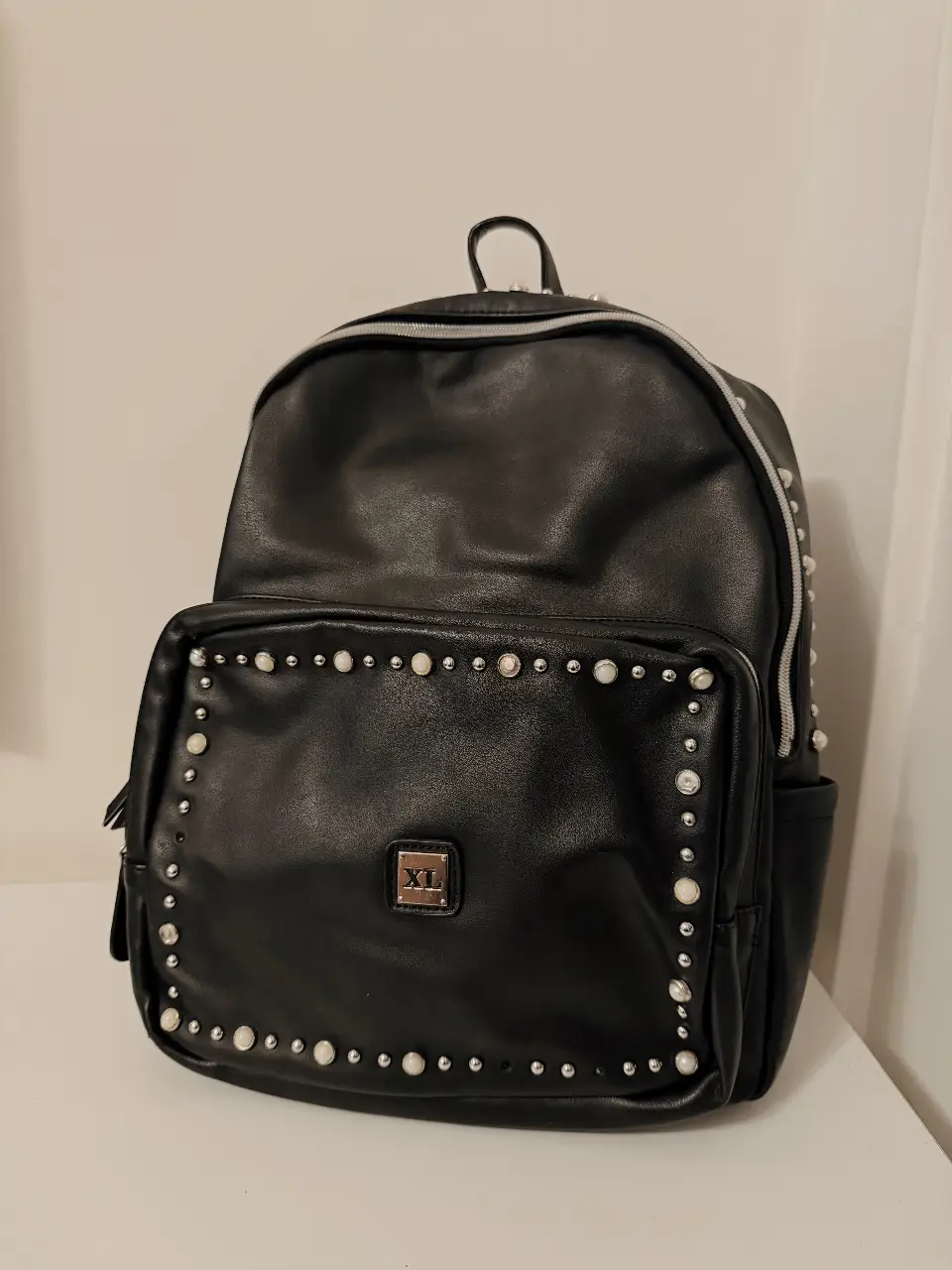 Mochila negra de ecocuero con detalles de tachas y perlas. Ideal para un look moderno y canchero. Cuenta con un bolsillo frontal con cierre y otro interno. Perfecta para llevar tus esenciales con estilo. detalle de uso en fotos