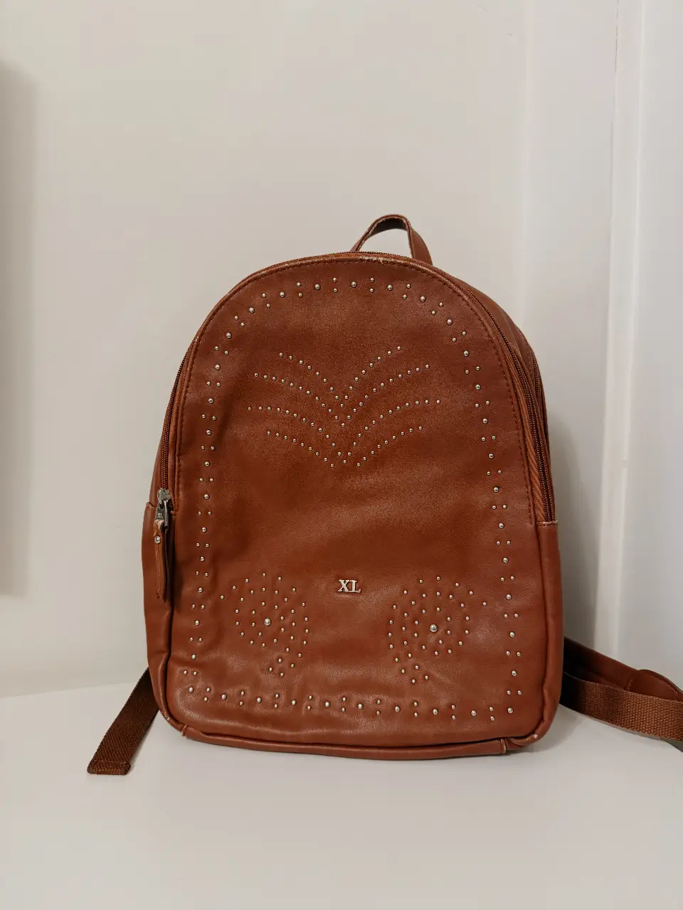 Mochila marrón con tachas plateadas, ideal para un look casual y canchero. Cuenta con un diseño con detalles de tachas en forma de "V" y dos círculos en la parte frontal. Tiene un compartimento principal con cierre, un bolsillo trasero con cierre y correas ajustables.