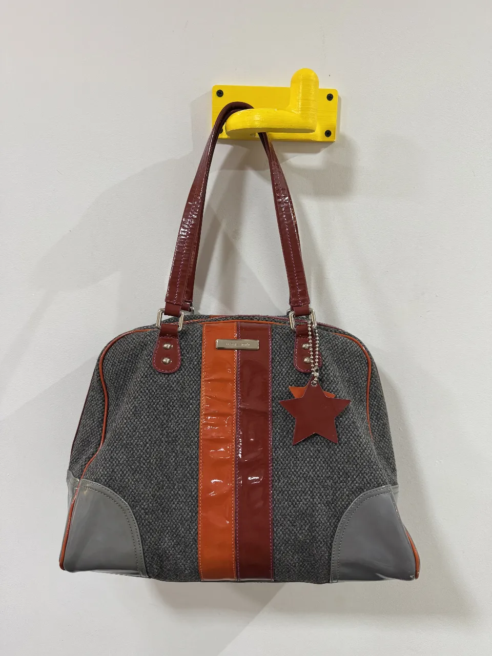 Cartera de mano para mujer, con diseño en tonos grises y detalles en naranja y marrón. Posee manijas superiores y una correa para colgar. Incluye un dije en forma de estrella.
